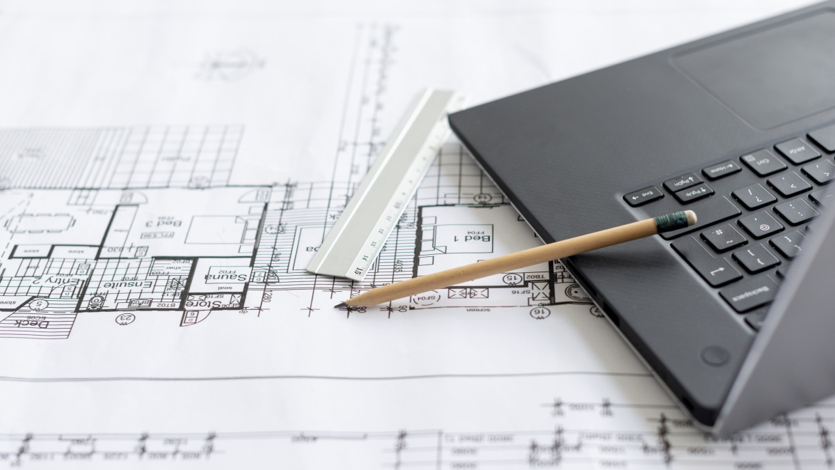 Un ordinateur portable, un plan de maison et des outils de tracage sont posés sur un bureau dans le but de réaliser une étude de structure. 
